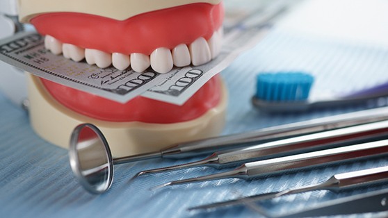 A model jaw biting hundred-dollar bills near dental instruments